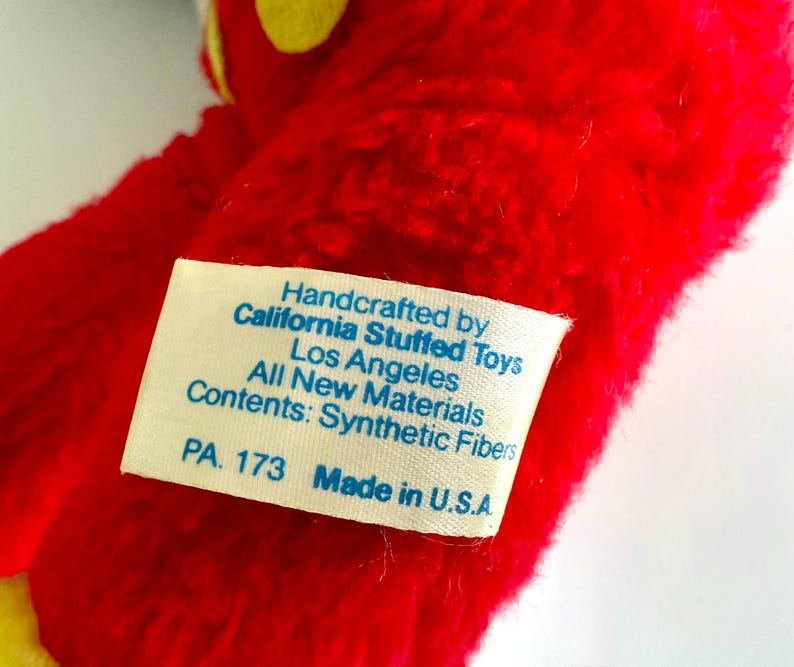 May include: Close-up of a red plush toy with yellow accents. A white tag is attached, displaying text: "Handcrafted by California Stuffed Toys, Los Angeles, All New Materials, Contents: Synthetic Fibers, PA. 173 Made in U.S.A."