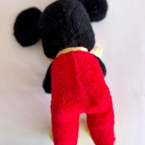 May include: A vintage plush toy of a cartoon mouse, viewed from the back. The toy has a black head with large, round ears, a red body, and yellow feet. A yellow collar is visible around the neck. The toy is made of a soft, plush material.