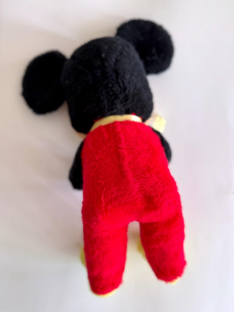May include: A vintage plush toy of a cartoon mouse, viewed from the back. The toy has a black head with large, round ears, a red body, and yellow feet. A yellow collar is visible around the neck. The toy is made of a soft, plush material.