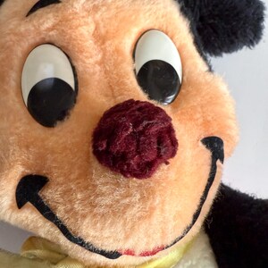 May include: Close-up of a vintage plush toy with a smiling face. The toy has a peach-colored face, large white eyes with black pupils, and a burgundy nose. It has black ears and a yellow ribbon around its neck.