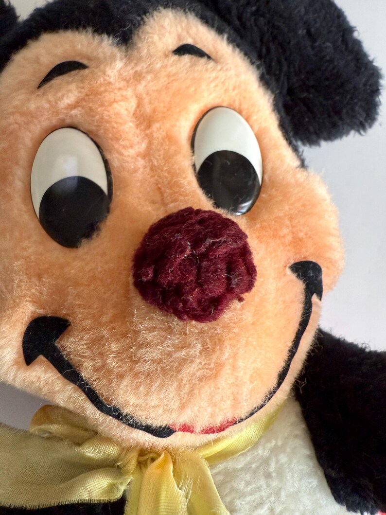 May include: Close-up of a vintage plush toy with a smiling face. The toy has a peach-colored face, large white eyes with black pupils, and a burgundy nose. It has black ears and a yellow ribbon around its neck.