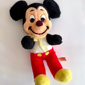 May include: A vintage Mickey Mouse plush toy. The plush features black ears, a tan face, and a red nose. Mickey wears red pants, yellow shoes, and a white shirt with yellow buttons and a yellow bow tie. A Walt Disney tag is attached.