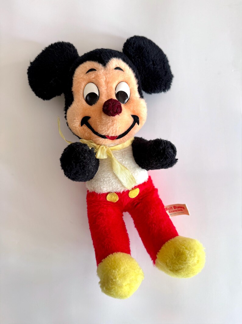 May include: A vintage Mickey Mouse plush toy. The plush features black ears, a tan face, and a red nose. Mickey wears red pants, yellow shoes, and a white shirt with yellow buttons and a yellow bow tie. A Walt Disney tag is attached.