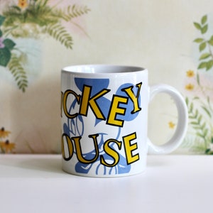 May include: White ceramic mug featuring the words "Mickey Mouse" in yellow outlined letters over a blue background. The mug has a white handle and is set against a floral and fauna background.