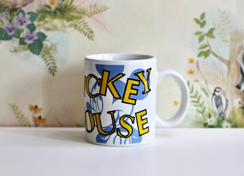 May include: White ceramic mug featuring the words "Mickey Mouse" in yellow outlined letters over a blue background. The mug has a white handle and is set against a floral and fauna background.