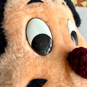 May include: Close-up of a vintage plush toy face with large, cartoonish eyes. The toy has a peach-colored face, black hair, and a burgundy nose. The eyes are white with black pupils. The toy appears to be made of soft, fuzzy material.