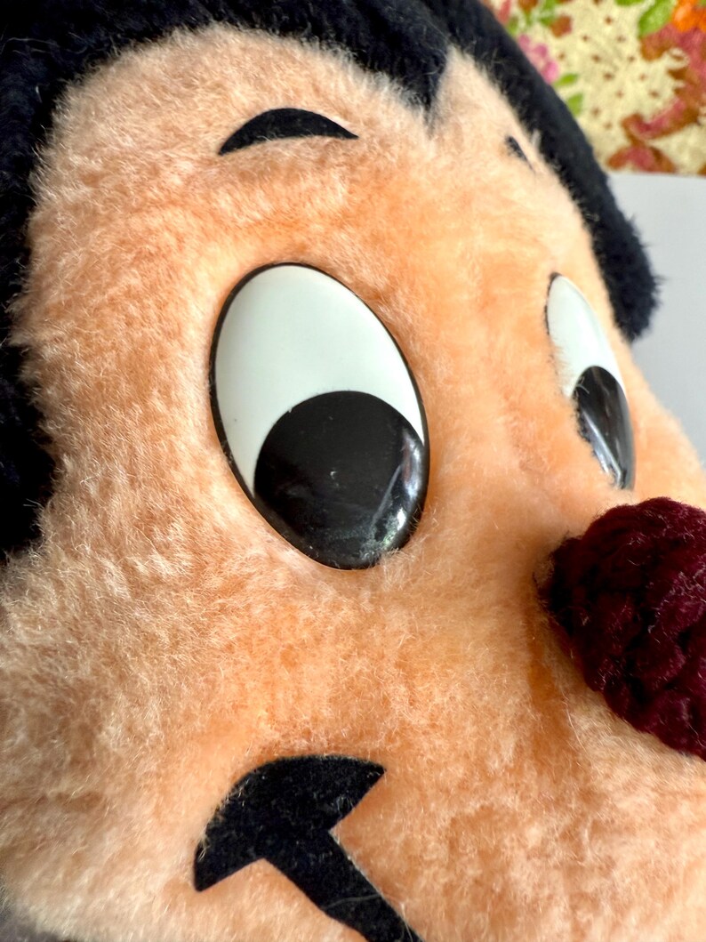 May include: Close-up of a vintage plush toy face with large, cartoonish eyes. The toy has a peach-colored face, black hair, and a burgundy nose. The eyes are white with black pupils. The toy appears to be made of soft, fuzzy material.