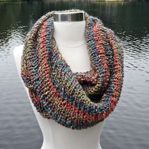 May include: A colorful knit infinity scarf with a mix of red, orange, green, blue, and purple yarn. The scarf is displayed on a white mannequin against a blurry background of a lake and trees.