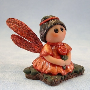 May include: A small, red-haired fairy figurine with a red dress and wings, holding a red rose. The fairy is sitting on a brown and green base.