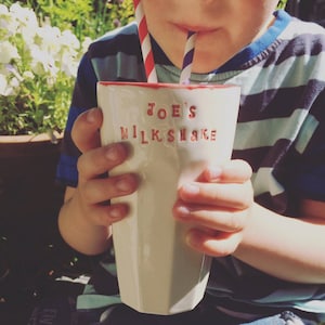 May include: A cream-coloured ceramic milkshake glass with the text "JOE'S MILKSHAKE" in red lettering. The rim of the glass is also red. Two straws, one red and white striped, the other purple and white striped, are in the glass.