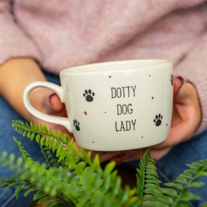 May include: Cream ceramic mug featuring the text "DOTTY DOG LADY" and black paw prints. The mug has a curved handle and small pink dots. The mug is held in someone's hands, with green foliage in the foreground.