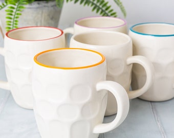 May include: Five white ceramic mugs with different coloured rims. The mugs have a textured, raised design and are perfect for a morning coffee or tea.