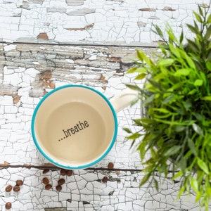 Peut inclure: Une tasse en céramique blanche avec un bord et une anse turquoise. La tasse porte l'inscription "...breathe" imprimée à l'intérieur. La tasse est posée sur une surface blanche craquelée avec des grains de café éparpillés autour.