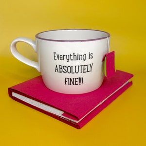 Può includere: Una tazza in ceramica bianca con una scritta nera che recita "Everything is ABSOLUTELY FINE!!!". La tazza è appoggiata su un quaderno rosa su uno sfondo giallo.