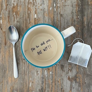 Puede incluir: Una taza de cerámica crema con borde y asa turquesa, con el texto "This too shall pass.... BUT WTF?!" Una cuchara plateada y una bolsita de té están junto a la taza sobre una superficie de madera.