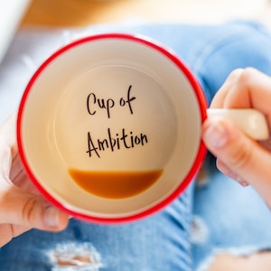 Puede incluir: Una taza de cerámica roja y blanca con las palabras "Cup of Ambition" escritas en el interior. La taza está llena de un líquido marrón.