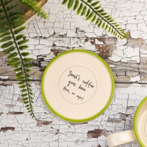 Puede incluir: Un posavasos de cerámica blanco con un borde verde. El posavasos tiene texto negro que dice "Sarah's coffee goes here (black, one sugar)"