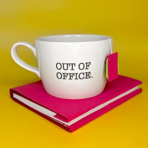 May include: White ceramic mug with a black inscription that reads "OUT OF OFFICE." The mug is sitting on top of a pink and white notebook.