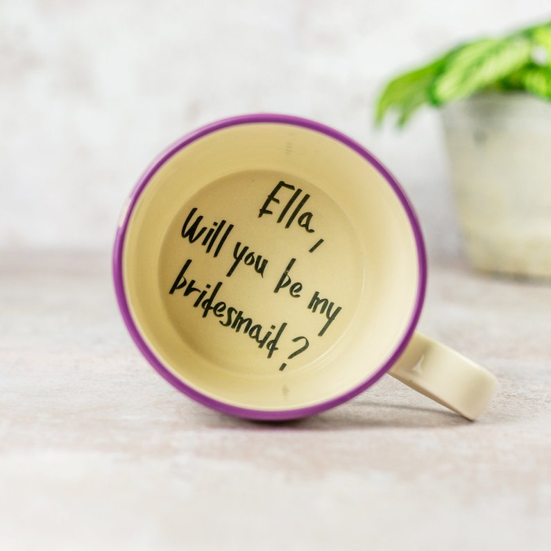 May include: Purple and white mug with the text "Ella, Will you be my bridesmaid?" written in black marker on the inside of the mug.