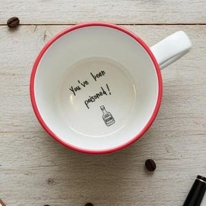 Peut inclure: Mug en céramique blanche avec un bord et une anse rouges. L'intérieur présente le texte "You've been poisoned!" et une petite illustration d'une bouteille. Des grains de café sont dispersés autour de la tasse sur une surface en bois.