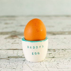 Op de afbeelding: Een bruin ei in een witte keramische eierdop met een turkooizen rand. De woorden "DADDY'S EGG" zijn in teal op de beker gedrukt. De achtergrond is een verweerd wit oppervlak.