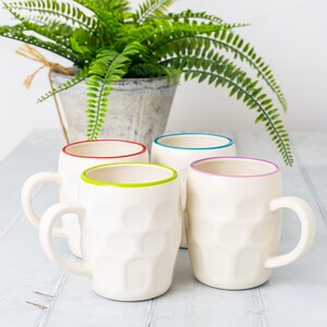 May include: Four white ceramic mugs with colored rims. The mugs have a textured, bumpy surface. The rims are red, blue, green, and purple.