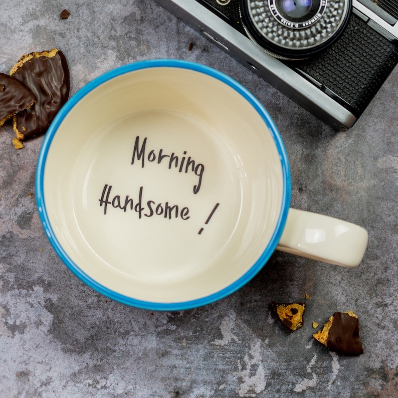 May include: A white ceramic mug with a blue rim and a handle. The mug has the words "Morning Handsome!" written inside.