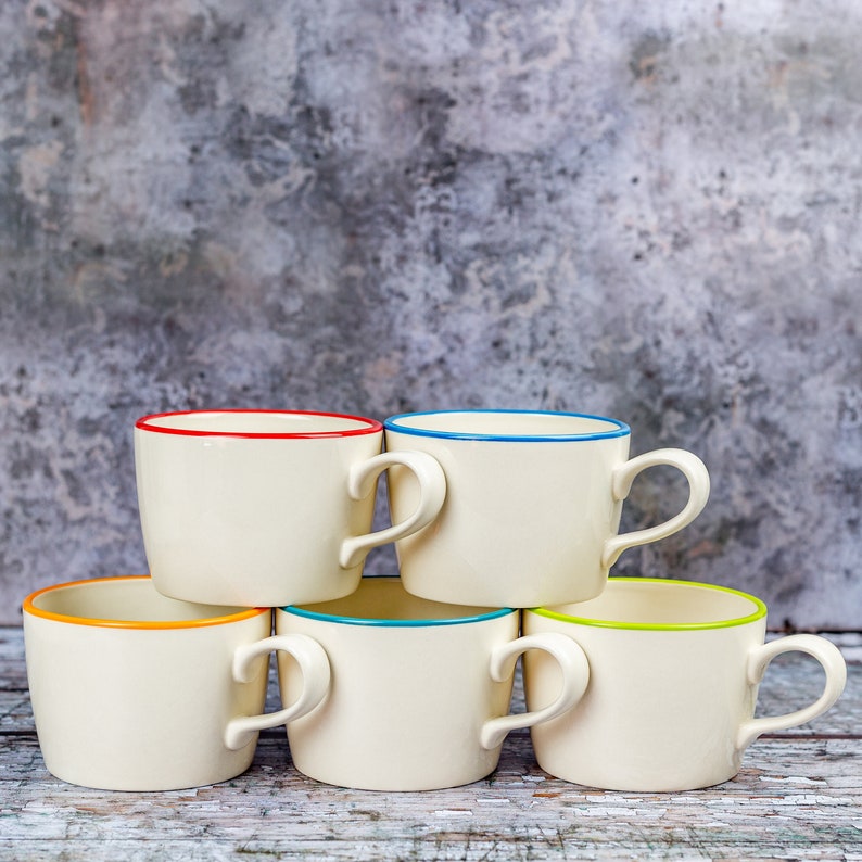May include: Four white ceramic mugs with colourful trim. The mugs are arranged in a row, with the handles facing outwards. The mugs are on a light grey background.