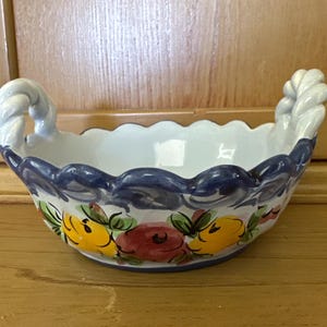 May include: A ceramic basket with a scalloped rim and rope-style handles. The basket is painted with a blue border and floral designs in yellow, red, and green. The interior is white.