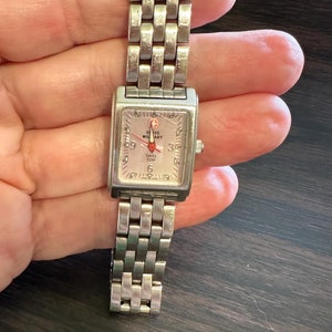 May include: A silver square-shaped watch with a white face and black numbers. The watch has a silver metal band and says "Swiss Military" on the face.