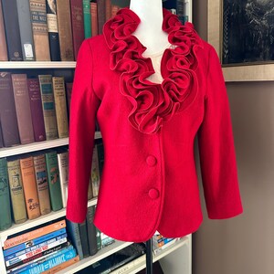 May include: A red wool blazer with a large, ruffled collar. The blazer has a single button fastening and long sleeves.