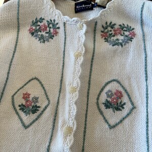 May include: White knitted cardigan with a crocheted trim and floral embroidery. The cardigan has a button closure and the label reads "Weekend Edition".
