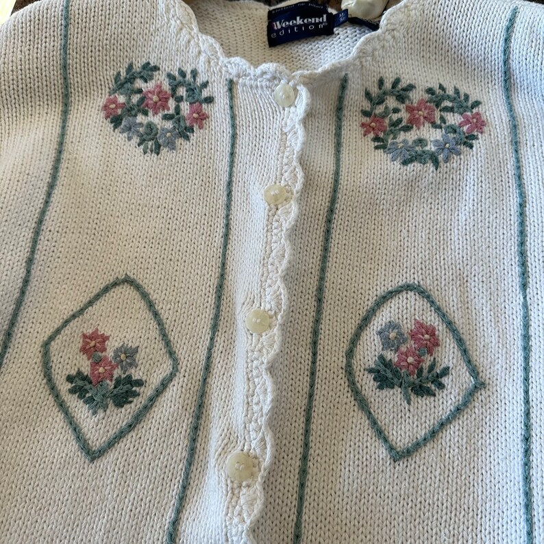 May include: White knitted cardigan with a crocheted trim and floral embroidery. The cardigan has a button closure and the label reads "Weekend Edition".