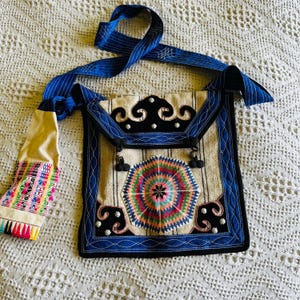 May include: A vintage embroidered fabric bag with a blue and white striped strap. The bag has a white background with a colorful circular design in the center and a black velvet trim. The bag is decorated with small black pom-poms.