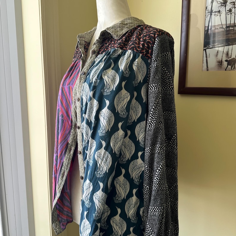 May include: A long-sleeved button-up shirt with a patchwork design. The shirt is made of different fabrics with various patterns, including a teal fabric with white bird silhouettes, a black fabric with white polka dots, and a purple and pink fabric with a wavy pattern.