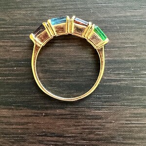 K&ouml;nnte beinhalten: Ein goldener Ring mit einer Reihe von bunten Edelsteinen. Die Edelsteine sind in einer goldenen Fassung gefasst und in einem Regenbogenmuster angeordnet. Der Ring liegt auf einer dunkelbraunen Holzoberfl&auml;che.