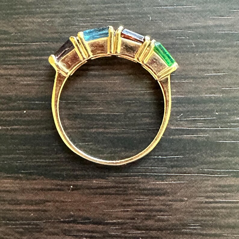 K&ouml;nnte beinhalten: Ein goldener Ring mit einer Reihe von bunten Edelsteinen. Die Edelsteine sind in einer goldenen Fassung gefasst und in einem Regenbogenmuster angeordnet. Der Ring liegt auf einer dunkelbraunen Holzoberfl&auml;che.