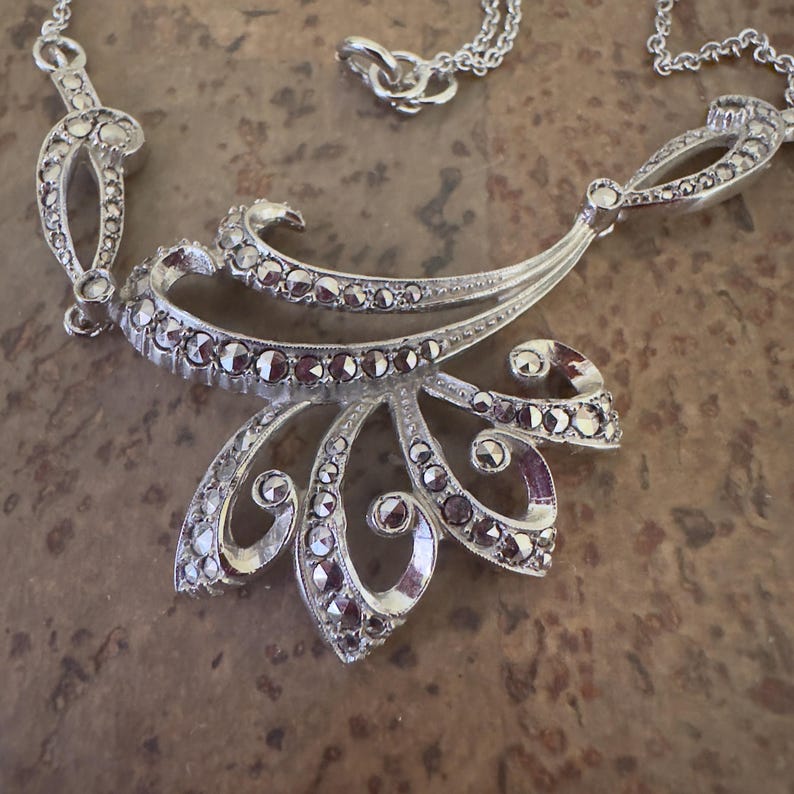 May include: Silver-toned necklace with a floral pendant. The pendant features a layered design with clear and dark-colored stones. The necklace has a delicate chain and a decorative clasp.