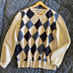 May include: A beige V-neck sweater with a classic argyle pattern in navy and light blue diamonds. The sweater has long sleeves and is displayed on a wooden hanger.