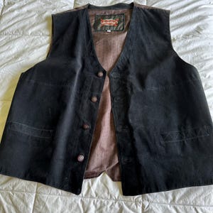 May include: A black suede waistcoat with a brown lining and multiple buttons. The waistcoat has two pockets on the front and a label on the inside that reads "Suede Leather".