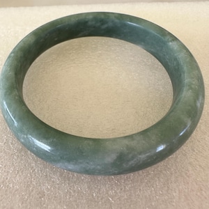 May include: A green jade bangle bracelet. The bangle is smooth and polished, with a light green color and white marbling.