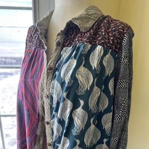 May include: A long-sleeved button-up shirt with a patchwork design. The shirt features a variety of fabrics and patterns, including a teal fabric with white bird silhouettes, a black and white polka dot fabric, and a pink and purple fabric with a swirling pattern.
