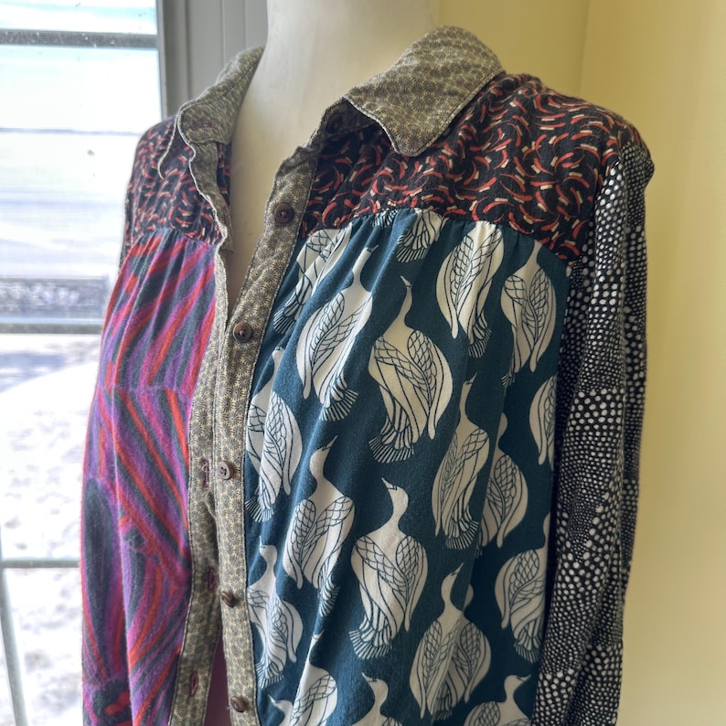 May include: A long-sleeved button-up shirt with a patchwork design. The shirt features a variety of fabrics and patterns, including a teal fabric with white bird silhouettes, a black and white polka dot fabric, and a pink and purple fabric with a swirling pattern.