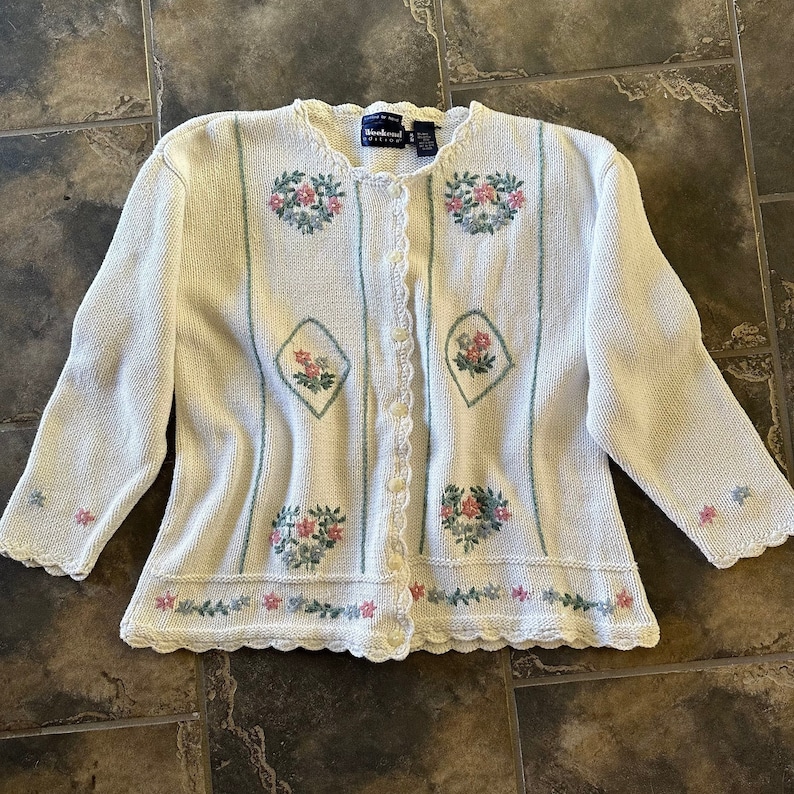 May include: A white crocheted cardigan with a scalloped hem and floral embroidery. The cardigan has a button-up front and is made of a soft, lightweight material.