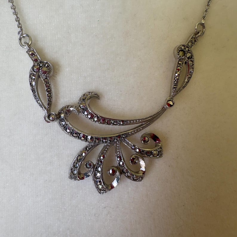 May include: A silver-toned necklace with a floral design. The necklace features a central, stylized flower with curved petals and a flowing, symmetrical design. Small, dark red stones are set throughout the piece.