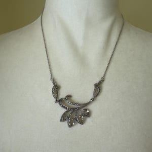 May include: A silver-toned necklace with a floral-inspired pendant. The necklace features a delicate chain and a detailed pendant with a leaf-like design, embellished with small, sparkling stones. The necklace is displayed on a mannequin.