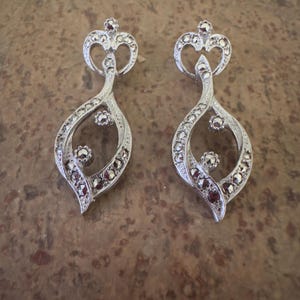 May include: A pair of silver-toned earrings with a crown-like top and a teardrop-shaped body. The earrings are adorned with small, dark-colored stones and have a vintage aesthetic. The design is intricate and detailed, suitable for formal occasions.