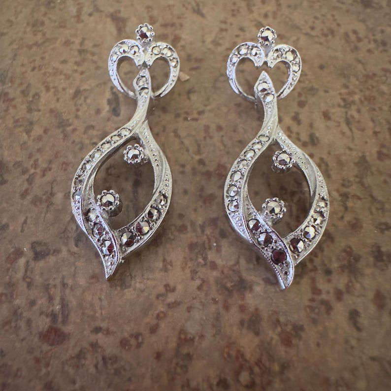 May include: A pair of silver-toned earrings with a crown-like top and a teardrop-shaped body. The earrings are adorned with small, dark-colored stones and have a vintage aesthetic. The design is intricate and detailed, suitable for formal occasions.