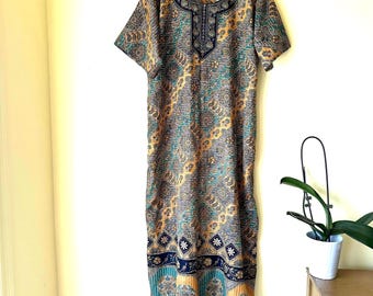May include: A floor-length kaftan dress featuring a multi-coloured paisley and geometric pattern in shades of gold, teal, and navy blue. The garment has short sleeves, a round neckline with a dark navy blue embroidered placket, and a wide hem.
