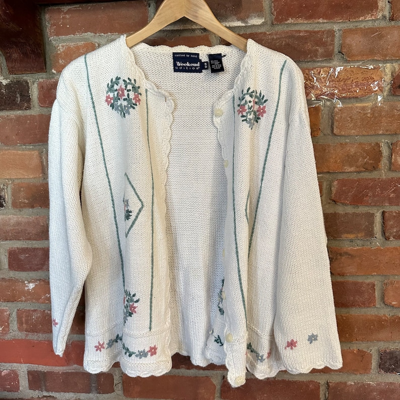 May include: A white knitted cardigan with green and pink floral embroidery. The cardigan has a scalloped hem and a button closure. The label reads "Weekend Quilts".
